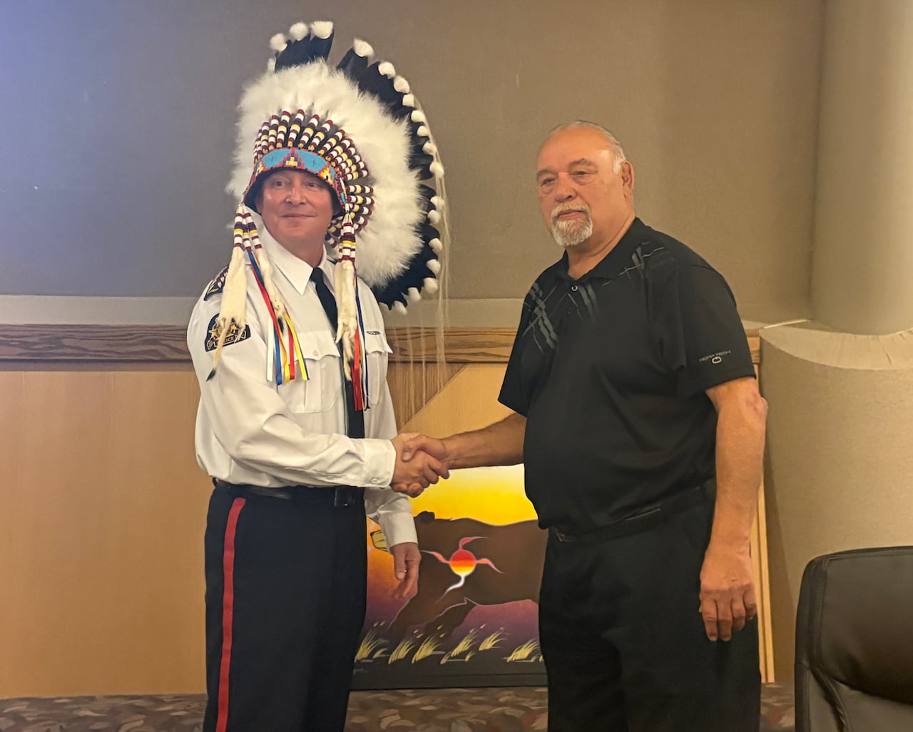 Two men stand beside each other shaking hands, one is is a uniform while wearing a feathered headress, the other has short hair and a light beard with dark clothing.